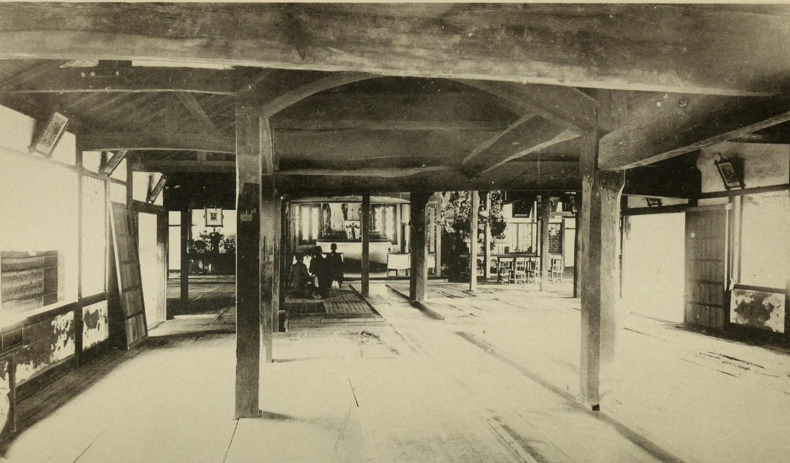 Photogravure of a bare wooden hall with heavy posts and beams, sliding screens open on both sides and a few people kneeling before a small altar at the far end