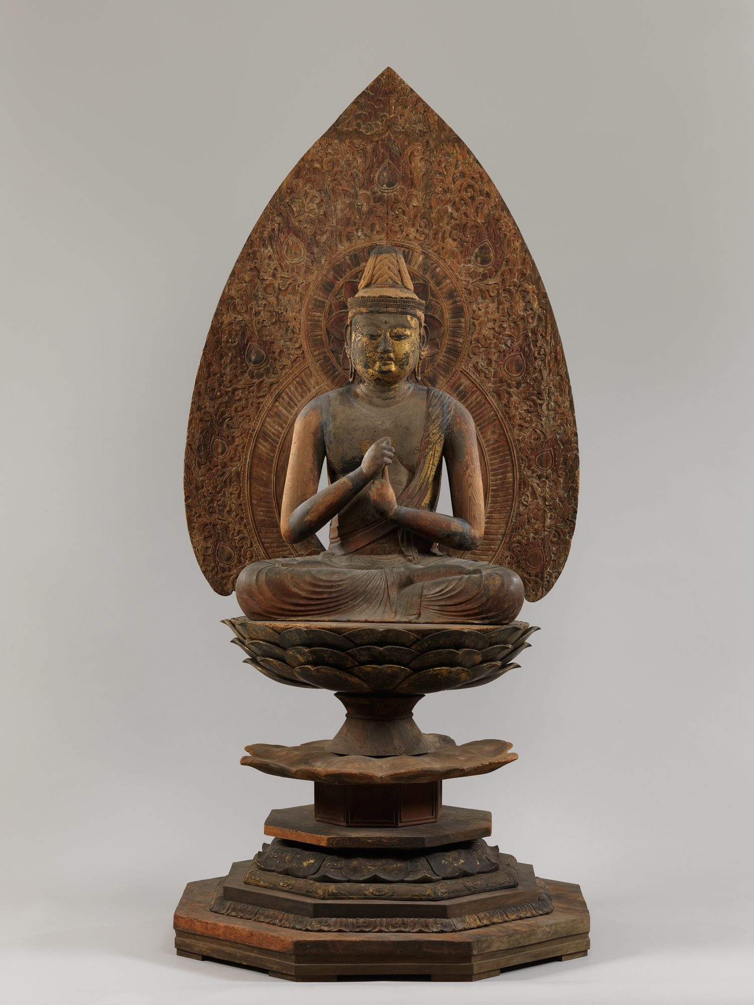 Carved wooden statue of a crowned Buddha seated on a tiered lotus pedestal, one hand clasping a raised finger of the other, before a flame-shaped halo
