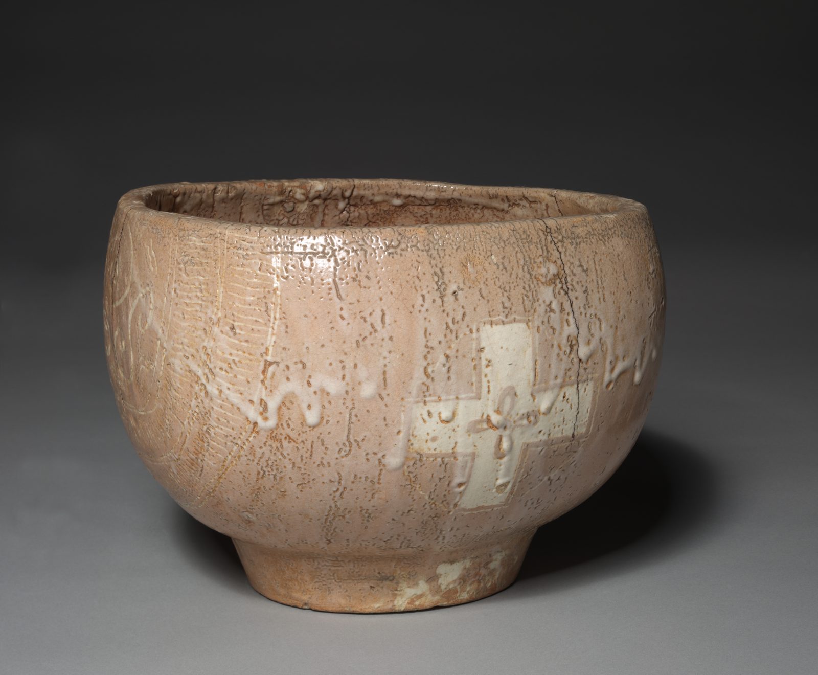 A deep stoneware bowl on a low foot, its pinkish crackled glaze streaked with white slip, with a large white cross painted on its side