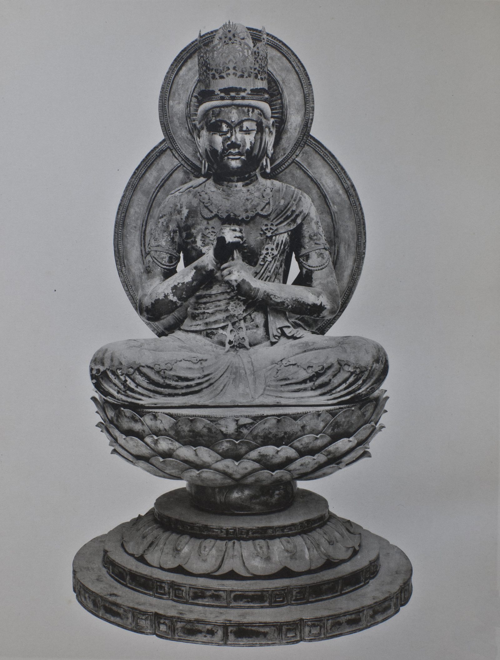 Black-and-white photograph of a crowned, bejeweled seated Buddhist statue with hands joined at the chest, before a double halo, on a lotus pedestal